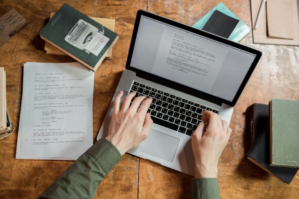 High-angle view of hands typing on a laptop surrounded by books and papers.